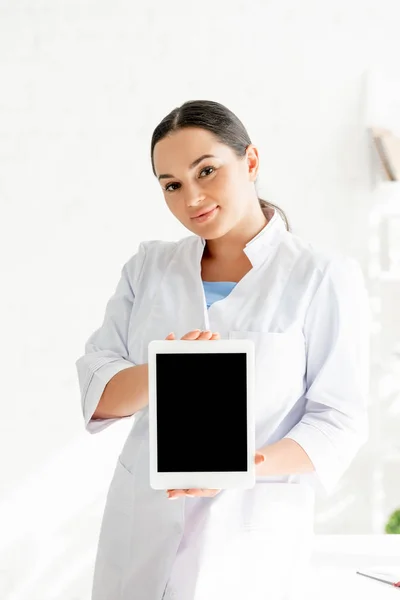 Atractivo dermatólogo sosteniendo tableta digital y mirando a la cámara en la clínica - foto de stock