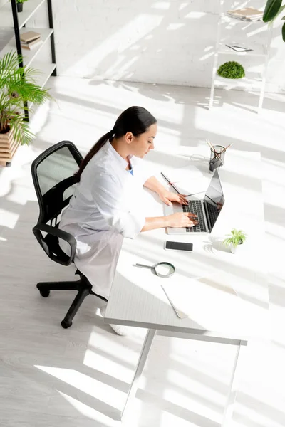 Vista ad alto angolo di dermatologo attraente seduto a tavola e utilizzando il computer portatile in clinica — Foto stock