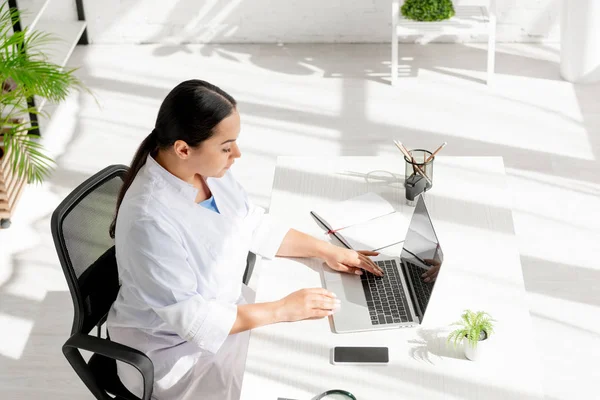 Blickwinkel auf attraktive Dermatologen, die am Tisch sitzen und Laptop in der Klinik benutzen — Stockfoto