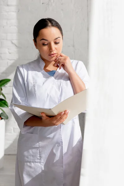 Selective focus of attractive dermatologist reading and holding folder in clinic — Stock Photo