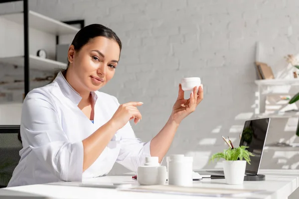 Atractivo dermatólogo sentado en la mesa y señalando con el dedo a la crema cosmética en la clínica - foto de stock