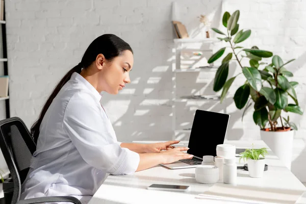 Attractive dermatologist sitting at table and using laptop in clinic — Stock Photo