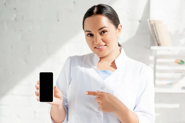 Lächelnder Hautarzt zeigt mit Finger auf Smartphone in Klinik — Stockfoto