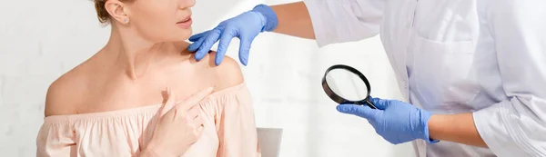 Panoramic shot of dermatologist examining skin of patient with magnifying glass in clinic — Stock Photo