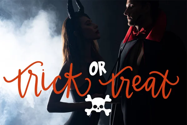 Handsome man in cloak looking at girl with horns holding flogging whip on black background with smoke and trick or treat illustration — Stock Photo