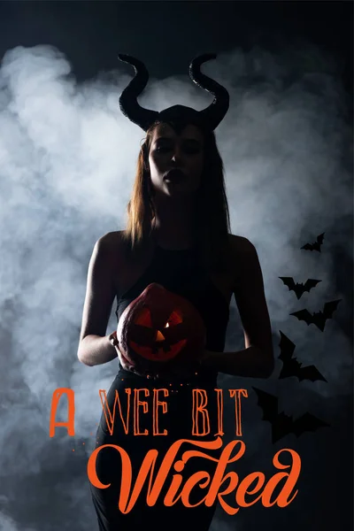 Silhouette of woman with horns holding spooky pumpkin on black background with smoke and a wee bit wicked illustration — Stock Photo