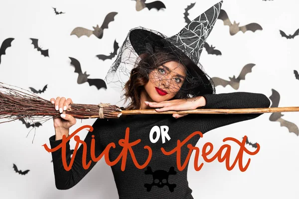 Attractive woman in witch hat holding broom in Halloween near trick or treat lettering — Stock Photo