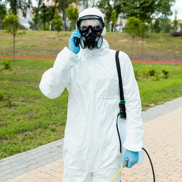 Reinigungsspezialist in Schutzanzug und Atemschutzmaske hält Sprühbeutel mit Desinfektionsmittel in der Hand, während er im Freien während der Coronavirus-Pandemie telefoniert — Stockfoto