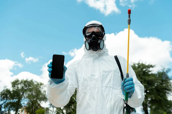 Especialista em limpeza em terno hazmat e respirador segurando saco de pulverização com desinfetante enquanto mostra smartphone com tela em branco ao ar livre durante a pandemia de coronavírus — Fotografia de Stock