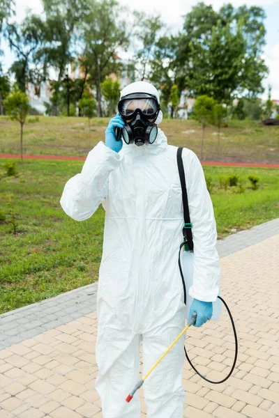 Especialista em limpeza em terno hazmat e respirador segurando saco de pulverização com desinfetante enquanto conversa no celular no parque durante a pandemia de coronavírus — Fotografia de Stock