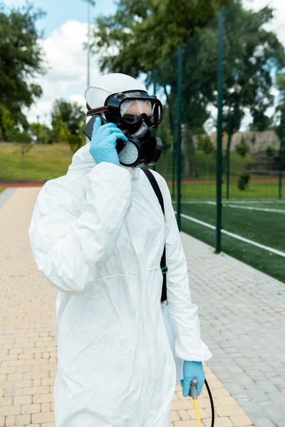 Cleaning specialist in hazmat suit and respirator holding spray bag with disinfectant while talking on cellphone outdoors during coronavirus pandemic — Stock Photo