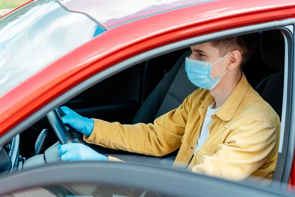 Männlicher Fahrer mit medizinischer Maske und Schutzhandschuhen, der während der Coronavirus-Pandemie das Lenkrad im Auto hält — Stockfoto