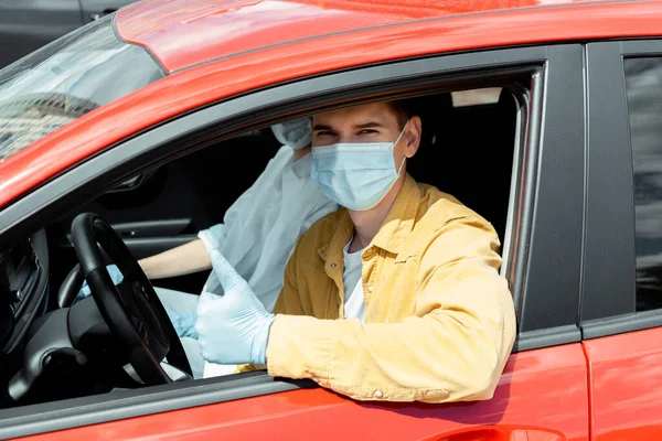 Man and woman in medical masks and protection gloves sitting in car and showing thumb up during covid-19 pandemic — Stock Photo