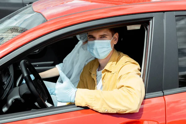 Homem e mulher em máscaras médicas e luvas de proteção sentados no carro e mostrando o polegar para cima durante a pandemia covid-19 — Fotografia de Stock