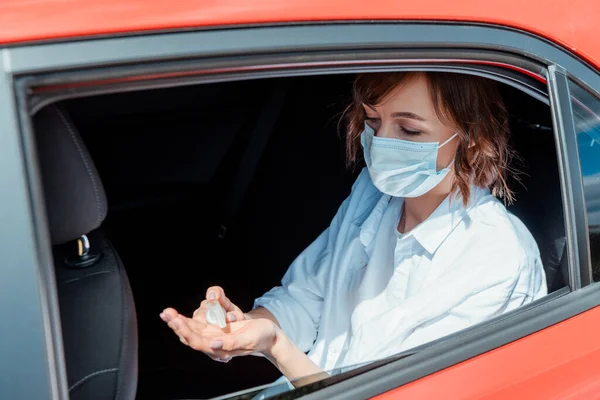Chica en máscara médica usando antiséptico en el coche durante la pandemia de coronavirus - foto de stock