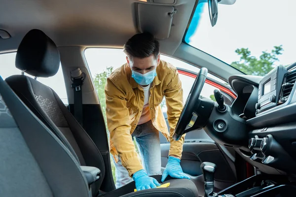 Uomo in maschera medica e guanti in lattice pulizia interni auto con straccio durante covid-19 pandemia — Foto stock