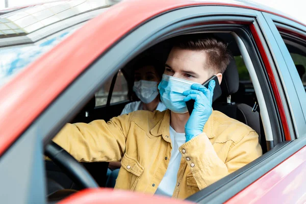 Male driver in medical mask and gloves talking on phone in taxi with passenger during coronavirus pandemic — Stock Photo