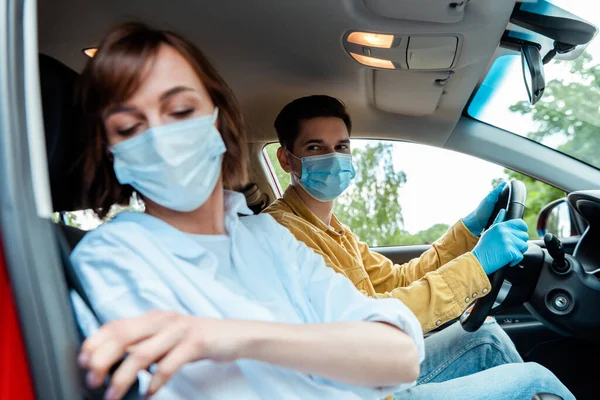 Uomo e donna in maschere mediche e guanti protettivi seduti in auto durante la pandemia coronavirica, focus selettivo — Foto stock