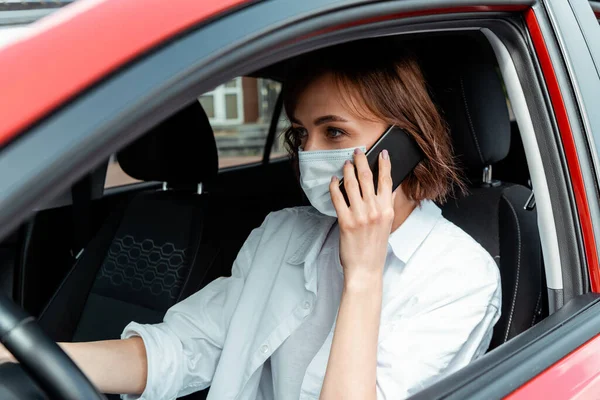 Женщина в медицинской маске разговаривает по мобильному телефону во время вождения автомобиля во время пандемии ковид-19 — стоковое фото