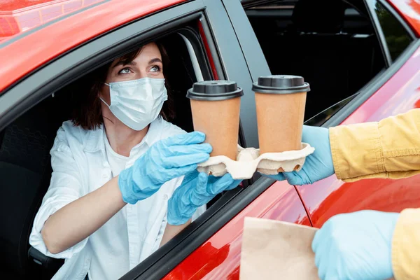 Femme en masque médical et gants acheter du café pour aller de la voiture pendant la pandémie de coronavirus — Photo de stock