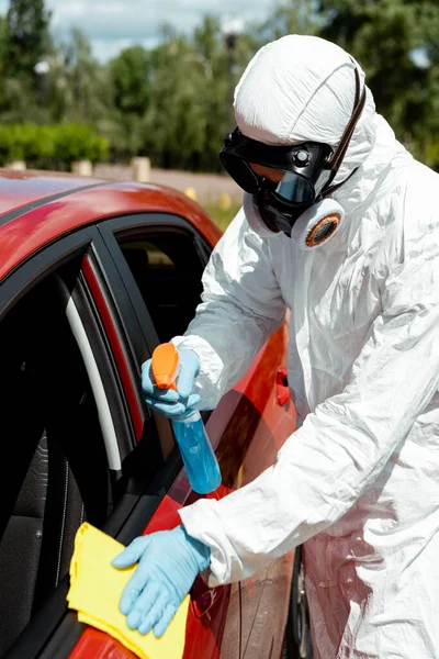 Especialista en limpieza de trajes de materiales peligrosos coche con spray antiséptico y trapo durante la pandemia de covid-19 - foto de stock