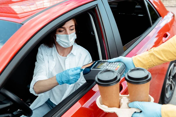 Woman in medical mask and gloves making transaction with credit card and terminal to buy takeaway coffee from car during covid-19 pandemic — Stock Photo