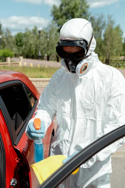 Cleaner in hazmat suit cleaning car with antiseptic spray and rag during coronavirus pandemic — Stock Photo