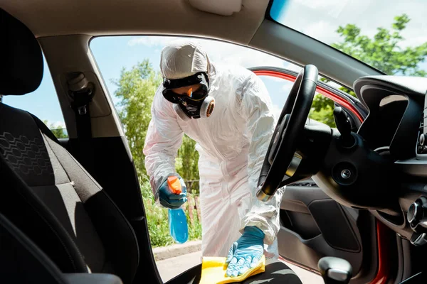 Especialista en traje de materiales peligrosos y respirador limpieza interior del coche con spray antiséptico y trapo durante la pandemia de coronavirus - foto de stock