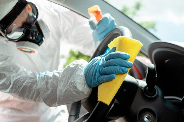 Specialist in hazmat suit and respirator cleaning car interior with antiseptic spray and rag during covid-19 pandemic — Stock Photo