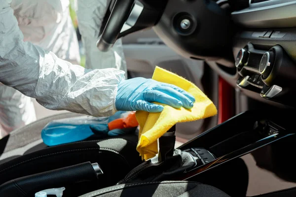 Обрезанный вид специалиста по чистке салона автомобиля защитного костюма с распылением и тряпкой во время пандемии коронавируса — стоковое фото