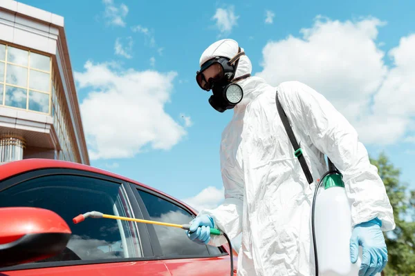 Workman in uniform and respirator cleaning car with disinfectant in spray bag during covid-19 pandemic — Stock Photo