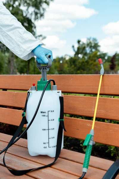 Vista recortada del especialista en traje de materiales peligrosos con desinfectante en bolsa de spray en el banco en el parque durante la pandemia de coronavirus - foto de stock