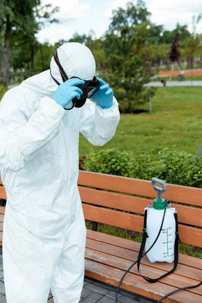 Specialist in hazmat suit and respirator disinfecting bench in park during coronavirus pandemic — Stock Photo