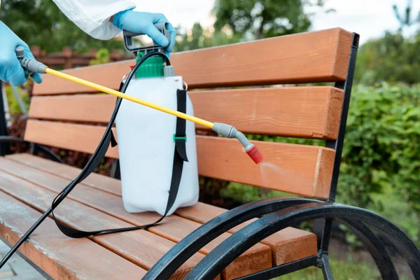 Cropped view of specialist in hazmat suit and respirator disinfecting bench in park during covid-19 pandemic — Stock Photo