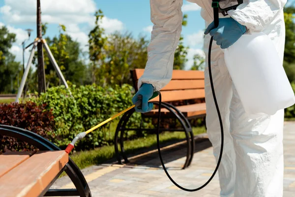 Specialist in hazmat suit and respirator disinfecting bench in park during coronavirus pandemic — Stock Photo