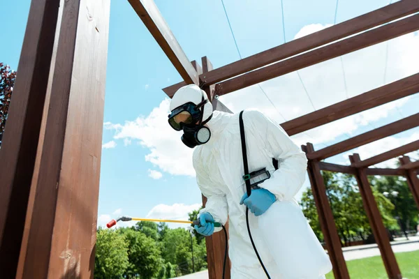 Specialist in hazmat suit and respirator disinfecting wooden construction in park during coronavirus pandemic — Stock Photo