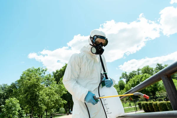 Especialista en traje de felpudo y respirador desinfectando barandillas en parque durante pandemia de covid-19 - foto de stock