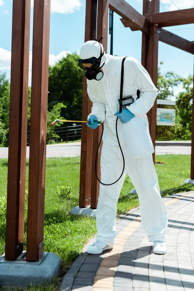 Professional specialist in hazmat suit and respirator disinfecting wooden construction in park during coronavirus pandemic — Stock Photo