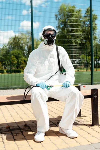 Especialista en traje de materiales peligrosos y respirador sentado en el banco con bolsa de spray durante la pandemia de covid-19 - foto de stock