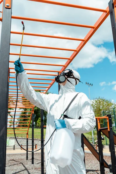 Trabajador en traje de felpudo y respirador desinfectando campo deportivo en parque durante pandemia de covid-19 - foto de stock