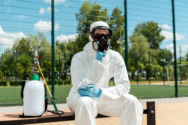 Pulizia specialista in hazmat tuta e respiratore pensiero e seduta su banco con sacchetto spray durante covid-19 pandemia — Foto stock