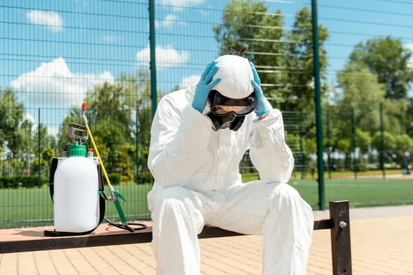 Especialista em limpeza estressado em terno hazmat e respirador sentado no banco com saco de pulverização durante a pandemia covid-19 — Fotografia de Stock