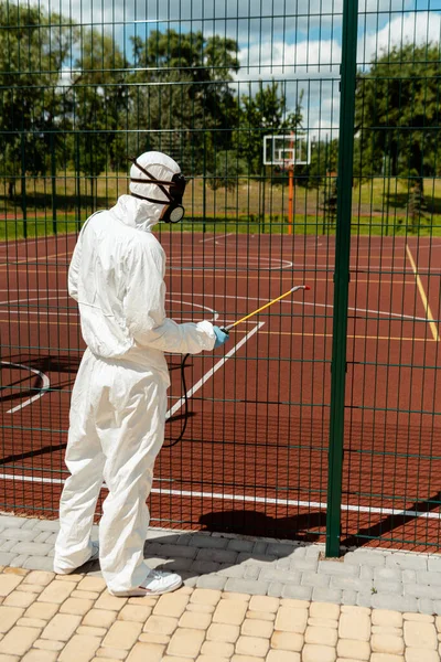 Especialista en traje de materiales peligrosos y respirador desinfectando cerca de cancha de baloncesto en parque durante pandemia de covid-19 - foto de stock