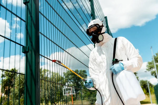 Male specialist in hazmat suit and respirator disinfecting fence of basketball court in park during covid-19 pandemic — Stock Photo