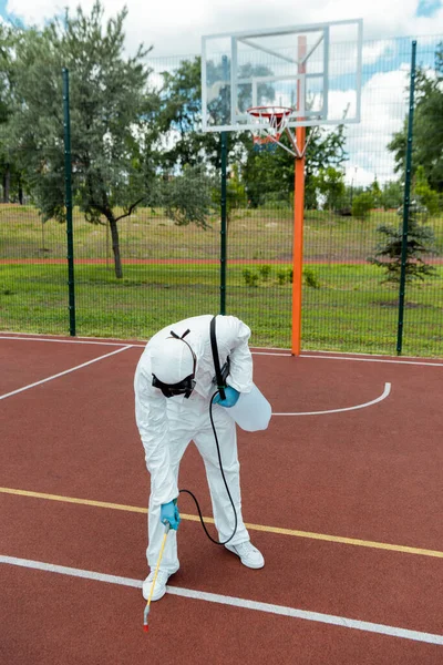 Specialist in hazmat suit and respirator disinfecting basketball court in park during covid-19 pandemic — Stock Photo