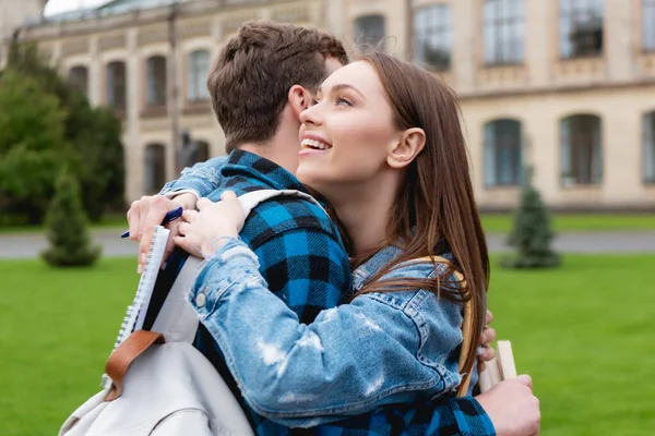 Atraente e feliz estudante abraçando amigo e segurando notebook com caneta — Fotografia de Stock