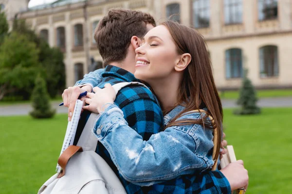 Studente attraente e felice con gli occhi chiusi abbracciando amico e tenendo il taccuino con la penna — Stock Photo