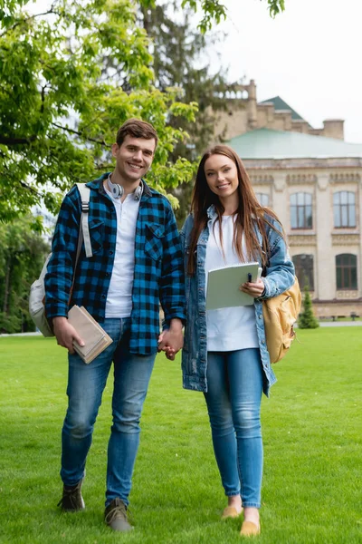 Cheerful students holding hands while standing on grass near university campus — Stock Photo