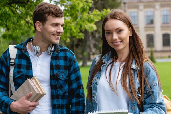 Étudiant gai avec écouteurs sans fil et des livres à la recherche de fille attrayante — Photo de stock