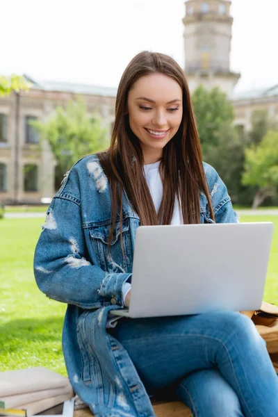 Estudiante alegre sentado en el banco y usando el ordenador portátil cerca de libros, concepto de estudio en línea - foto de stock